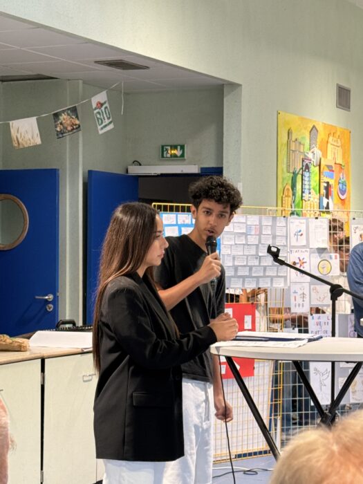 Cérémonie de transmission du drapeau Unadif au collège André-Malraux de La Farlède. Deux collégiens, Fouzi et Julia, maîtres de cérémonie, animent l’événement. Fouzi tient un micro et s’exprime devant le public, tandis que Julia se tient à ses côtés. En arrière-plan, une fresque colorée et des affiches décorent la salle, créant une ambiance solennelle et engageante."