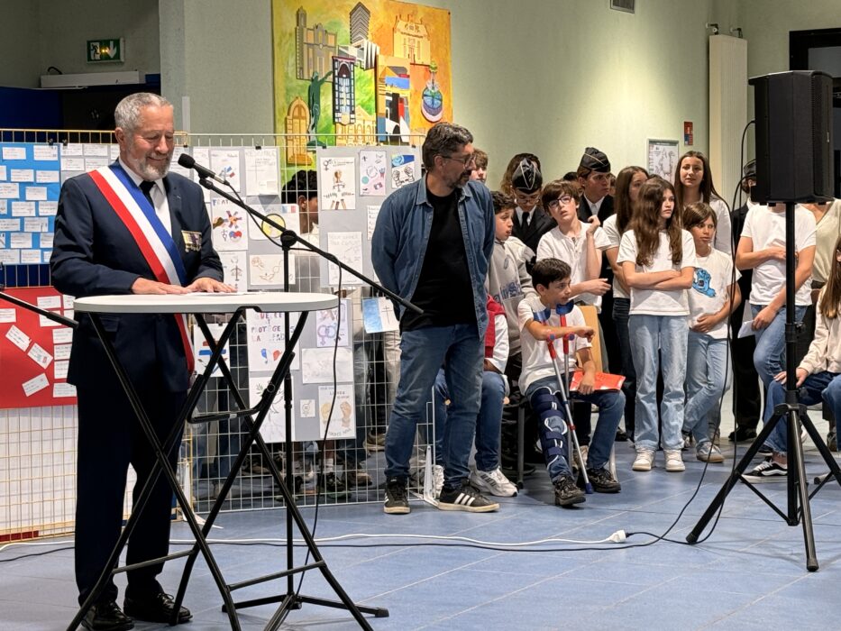 Cérémonie de transmission du drapeau Unadif au collège André-Malraux de La Farlède. Un élu portant l’écharpe tricolore s’exprime au micro devant un public composé d’élèves et d’adultes. Certains élèves, debout ou assis, portent des tenues formelles ou des bérets. En arrière-plan, une fresque colorée et des affiches illustrées sur les thèmes de la paix et de la mémoire décorent la salle.
