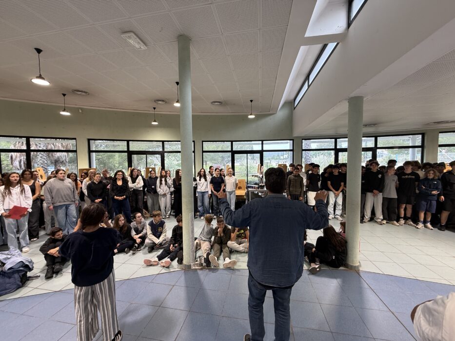 Cérémonie de transmission du drapeau Unadif au collège André-Malraux de La Farlède. Un groupe d’élèves et d’adultes est rassemblée dans une salle lumineuse avec de grandes baies vitrées. Certains sont debout, d’autres assis par terre, écoutant attentivement un animateur qui s’exprime au centre de la salle. L’ambiance est solennelle et collective.