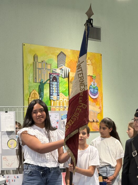 Cérémonie de transmission du drapeau Unadif au collège André-Malraux de La Farlède. Une collégienne, vêtue d’un t-shirt blanc et d’un jean, tient fièrement le drapeau des Internés de la Résistance, aux couleurs bleu, blanc, rouge avec l’inscription 'INTERNES DE LA RESISTANCE'. En arrière-plan, une fresque colorée représentant des bâtiments et des symboles de paix décore le mur, et d’autres élèves en t-shirt blanc assistent à la cérémonie.