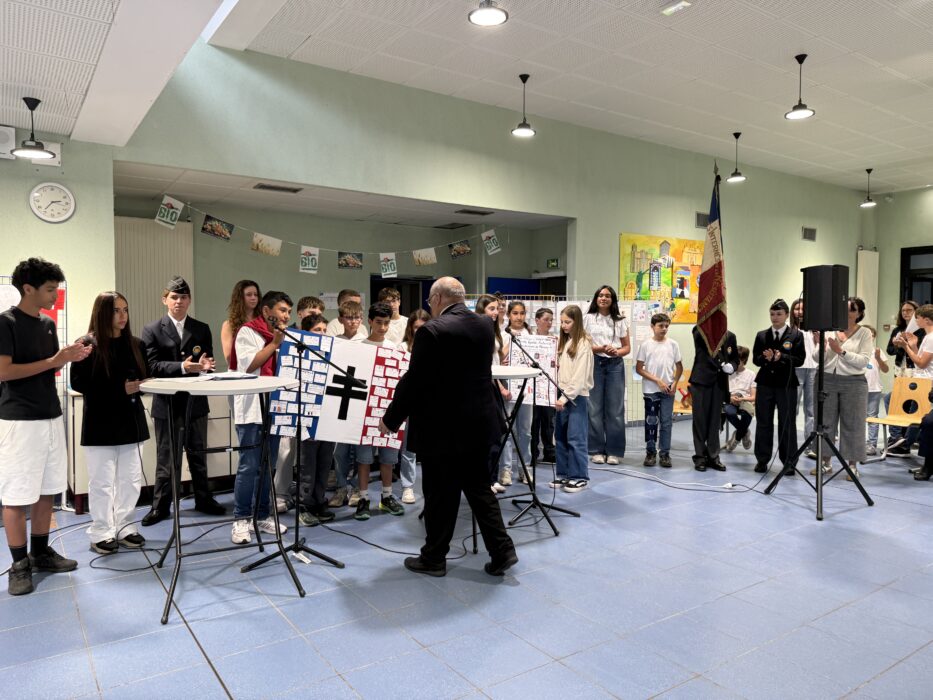 Cérémonie de transmission du drapeau Unadif au collège André-Malraux de La Farlède. Un homme en costume s’adresse à un groupe d’élèves et d’adultes réunis autour de pupitres équipés de microphones. Un panneau décoré aux couleurs de la France (bleu, blanc, rouge) avec une croix noire est visible au premier plan. Certains élèves portent des uniformes, et un drapeau français est accroché au mur en arrière-plan.