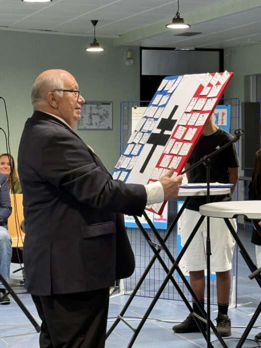 Cérémonie de transmission du drapeau Unadif au collège André-Malraux de La Farlède. Un homme en costume présente un panneau décoré aux couleurs de la France, sur lequel sont accrochées des cartes ou des messages. Il s’exprime devant un public assis, dans une salle équipée d’un micro et d’un pupitre. L’ambiance est solennelle et éducative.
