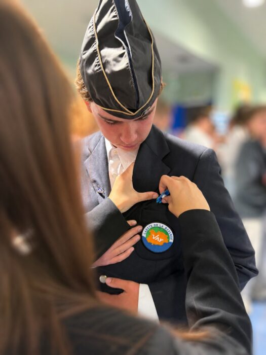 Cérémonie de transmission du drapeau Unadif au collège André-Malraux de La Farlède. Un élève, vêtu d’un costume sombre et d’un béret noir orné d’une bordure dorée, reçoit une épingle ou une médaille. Sur sa veste, un insigne circulaire porte l’inscription 'Cadets de la Défense' avec le logo du Var. Une personne à gauche l’aide à ajuster sa distinction.