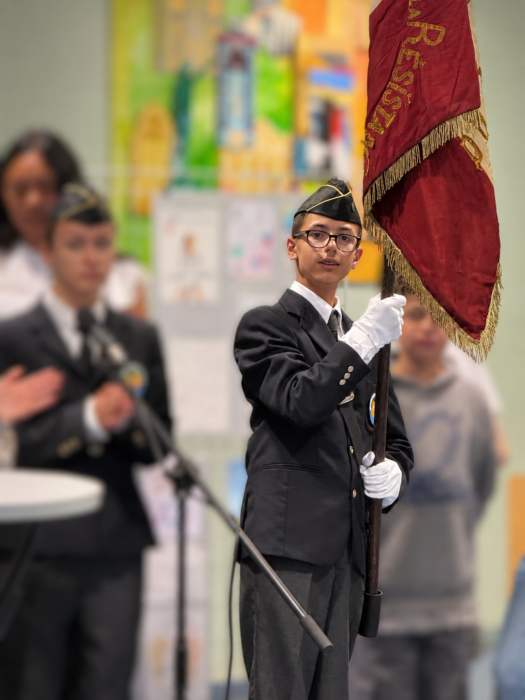 Cérémonie de transmission du drapeau Unadif au collège André-Malraux de La Farlède. Un élève, vêtu d’un costume sombre, d’une cravate, de gants blancs et d’un béret, porte fièrement un drapeau aux couleurs marron et or, avec des inscriptions dorées. Il tient le drapeau d’une main et le mât de l’autre, dans une posture solennelle. En arrière-plan, d’autres élèves en tenue formelle participent à la cérémonie.