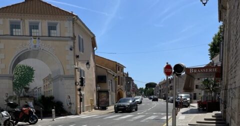 Rue urbaine ensoleillée avec passage piéton, voitures, bâtiments aux façades claires et arche décorée à l’entrée d’une ville du sud de la France.