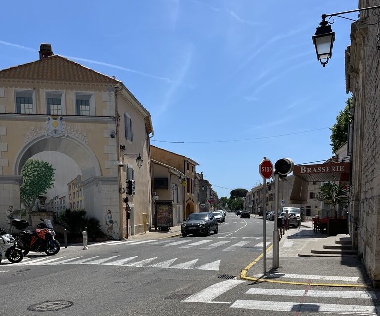 Rue urbaine ensoleillée avec passage piéton, voitures, bâtiments aux façades claires et arche décorée à l’entrée d’une ville du sud de la France.