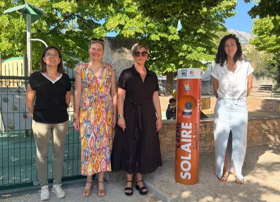 « Photo de l'inauguration de la Sun'Box dans le parc Gensollen à La Farlède. Quatre femmes posent devant un distributeur orange de crème solaire Sun'Box. Le distributeur porte les inscriptions 'Crème Solaire Sun'Box' et 'CPTS'. En arrière-plan, un parc avec des arbres, une aire de jeux pour enfants, et un mur en pierre. »