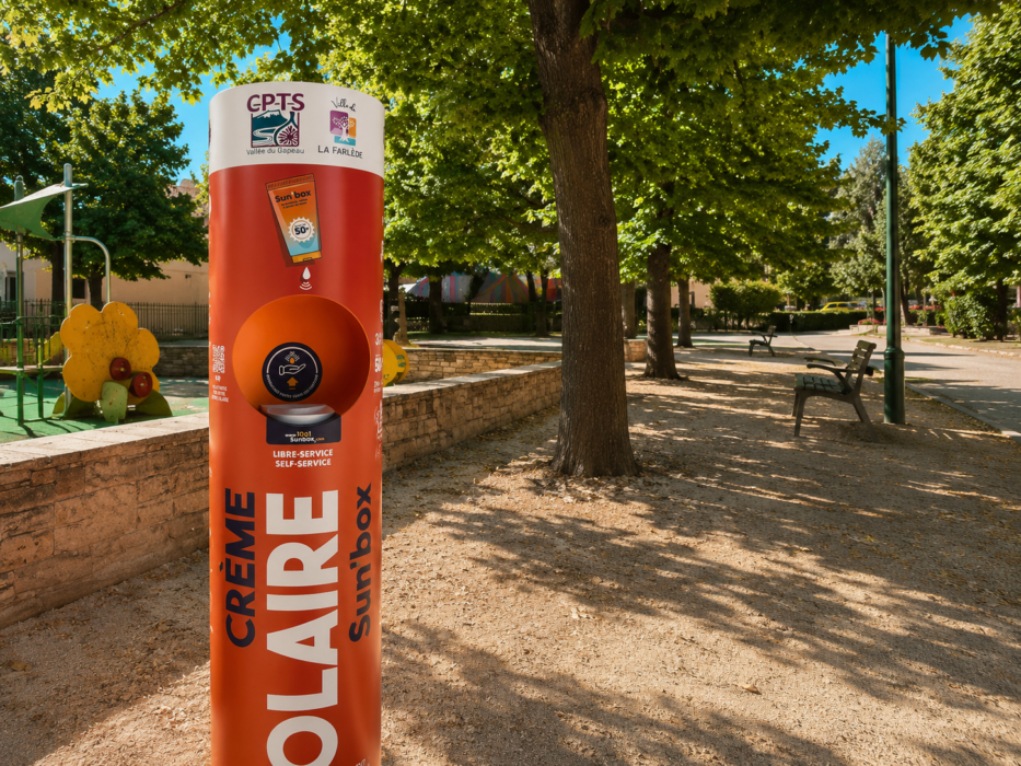 « Photo d'une Sun'Box (distributeur de crème solaire) installée dans le parc Gensollen à La Farlède. Le distributeur orange porte les inscriptions 'Crème Solaire Sun'Box', 'Libre-service / Self-service', 'CPTS Vallée du Gapeau' et 'La Farlède'. En arrière-plan, une aire de jeux pour enfants avec des structures colorées, des arbres, des bancs et un chemin en gravier. »