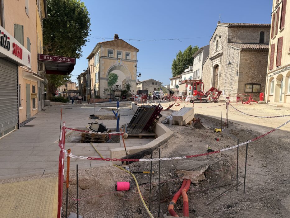 Chantier de travaux sur l’avenue de la République à La Farlède. Une tranchée ouverte au centre de la rue révèle des tuyaux orange et des câbles, protégée par des barrières et des chaînes rouges et blanches. Des matériaux de construction, comme des palettes en bois et des blocs de pierre, sont dispersés autour. En arrière-plan, des bâtiments historiques, dont une église, une brasserie et des maisons aux volets colorés, bordent la rue. Une pelleteuse rouge est visible, et des ouvriers travaillent sous un ciel bleu dégagé.
