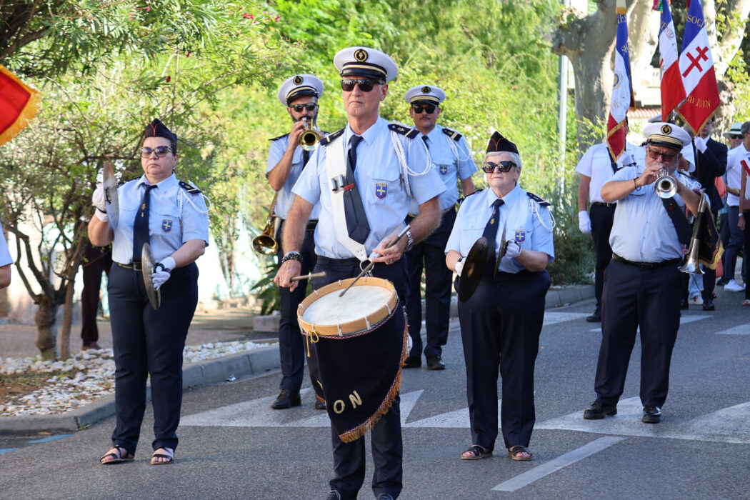 « Photo d'un défilé musical lors de la fête de la Libération à La Farlède, le 21 août 2026. Une fanfare en uniforme marche dans la rue, jouant des instruments comme des trompettes, des tambours et des cymbales. Les musiciens portent des tenues officielles avec casquettes, chemises blanches et pantalons noirs. Des drapeaux, dont le drapeau français, sont visibles en arrière-plan. »
