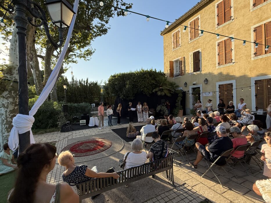 Soirée de lancement de 'Fêter le territoire' au jardin Pagès à La Farlède. Un public assis sur des chaises en plein air écoute des intervenants debout sur une scène improvisée. Les participants sont entourés d’arbres, de palmiers et de bâtiments, avec le soleil couchant créant une lumière dorée et des reflets de lens flare. L’ambiance est chaleureuse et conviviale, dans un cadre naturel et urbain.