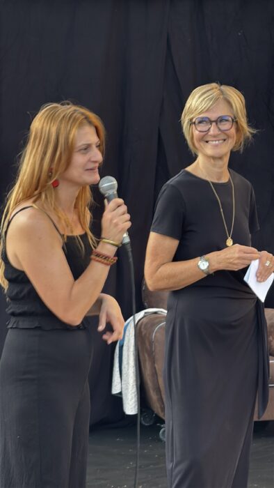 Soirée de lancement de 'Fêter le territoire' au jardin Pagès à La Farlède. À gauche, Christelle Didero, co-responsable de la médiathèque Eurêka, tient un micro et s’exprime avec engagement. À droite, Anne-Laure Excoffon-Jolly, adjointe à la culture, écoute attentivement en tenant des notes. Les deux femmes sont habillées en noir, sous un fond de scène sombre, créant une ambiance sérieuse et dédiée à l’événement.