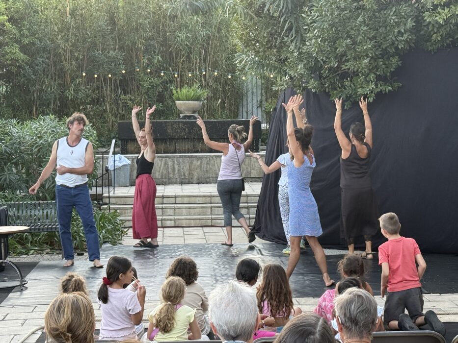 Spectacle de théâtre participatif lors de la soirée de lancement de 'Fêter le territoire' au jardin Pagès à La Farlède. Cinq artistes, dont un homme en débardeur blanc et jean, et quatre femmes en tenues colorées, animent une scène en plein air devant un public assis, composé d’enfants et d’adultes. En arrière-plan, une haie de bambous et des guirlandes lumineuses créent une ambiance chaleureuse et festive.