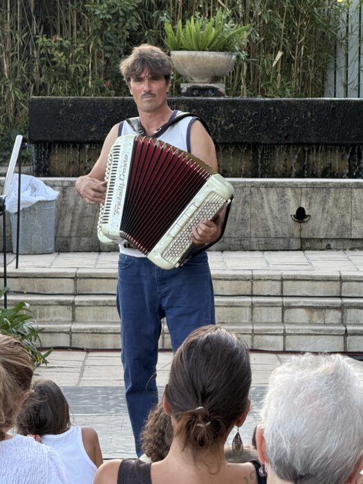Animation musicale lors de la soirée de lancement de 'Fêter le territoire' au jardin Pagès à La Farlède. Un musicien joue de l’accordéon sur une scène en plein air, devant un public assis composé d’enfants et d’adultes. En arrière-plan, une haie de bambous, une fontaine en pierre et des plantes vertes créent une ambiance naturelle et conviviale.